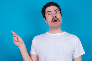 Emotive young handsome Caucasian man with moustache wearing white  keeps jaw dropped from shock demonstrates amazing promo points right on blank space demonstrates big shopping sale. Advertisement