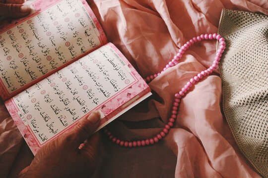 womans hands holding a copy of Quran, book of Islam religion, papers with prayers in Arabic language,  with pink rosary and taqiyah in pink textile background 