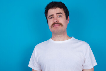 Fototapeta premium Displeased young handsome Caucasian man with moustache wearing white t-shirt against blue background frowns face feels unhappy has some problems. Negative emotions and feelings concept
