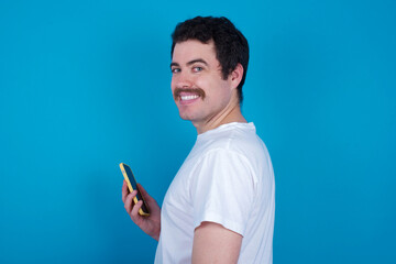 Rear view photo portrait of young handsome Caucasian man with moustache wearing white t-shirt against white background using smartphone smiling