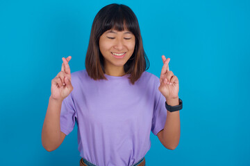 Young beautiful asian girl wearing purple t-shirt against blue background has big hope, crosses fingers, believes in good fortune, smiles broadly. People and wish concept