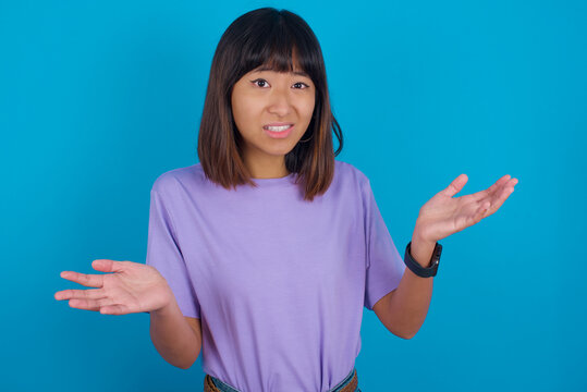 Clueless Young beautiful asian girl wearing purple t-shirt against blue background shrugs shoulders with hesitation, faces doubtful situation, spreads palms, Hard decision
