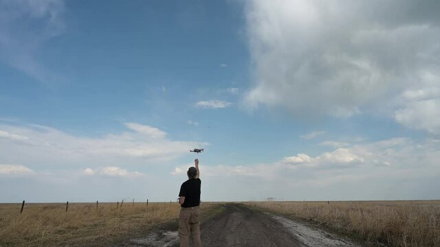 Man in a black shirt having difficultly in catching a landing drone with a blue sky and white clouds in the background.
