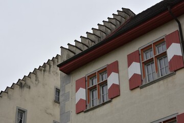 Castle in city Tiengen, Germany. Detail of facade with two windows and shutters in red and white colors and articulated roof edges. Photo made in low angle view. There is overcast sky on background. 