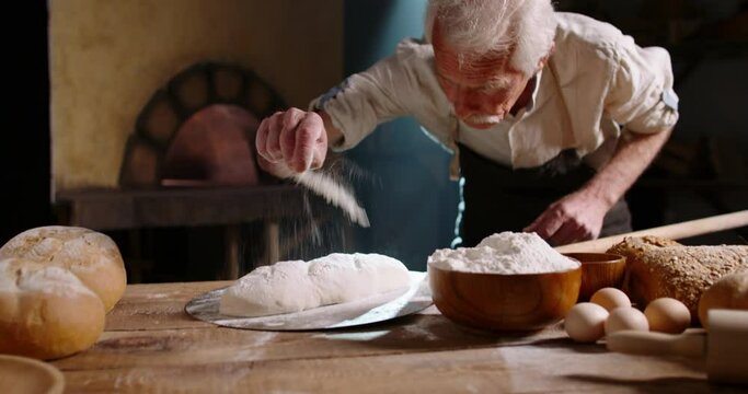 Senior bakery worker putting flour on bread dough and forming a loaf, putting the bun into stone oven - meal preparation, food and drink 4k footage