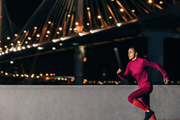 Woman running at night on embankment. Side view of female jogger sprinting outdoors against bridge.