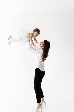 Happy harmonious family indoors in studio. Attractive young mother wearing high heels and white blouse throws the baby girl up, laughs and plays together on the white background