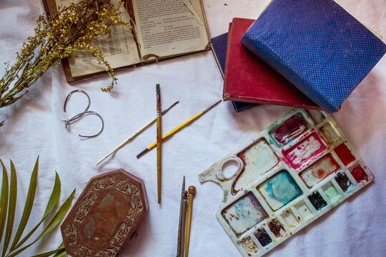 White bedsheet with art supplies and leather-bound journals on it around leaves and a jewelry box