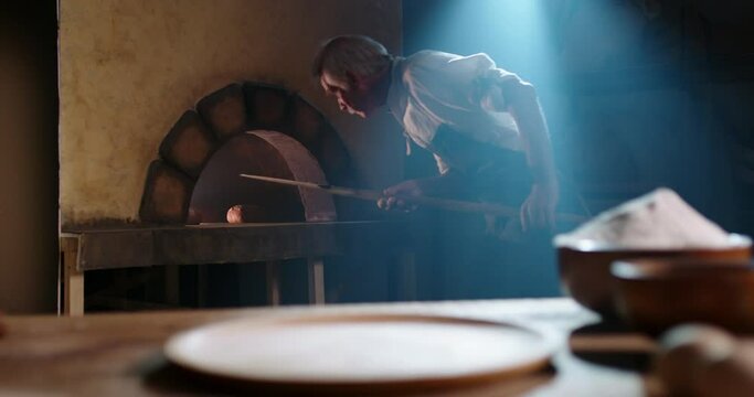 Senior bakery worker taking a peel to take cooked hot bread loaves out of stone oven and putting bun on table - food and drink, food processing, meal preparation 4k footage