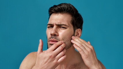 Fototapeta premium Portrait of shirtless handsome brunette guy checking his skin, looking anxious, posing isolated over blue background