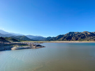Fototapeta premium Lake and mountains landscape on clear day in Potrerillos, Mendoza. Distant view of the high mountains and the people next to the water in summer.
