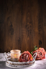 Drei Rosen in Alt Rose' Farben und ein Kerzenständer mit Perle liegen auf dem Holzhintergrund auf dem weißen Tablett aus Metall. Vintage, Hochformat, Hintergrund. Oben viel Platz zum beschriften.