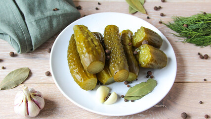 Plate of pickled homemade cucumbers on wooden rustic background. Marinated pickled cucumbers on the white plate with garlic, black pepper and bay leaf. Food concept. Homemade pickled vegetables.