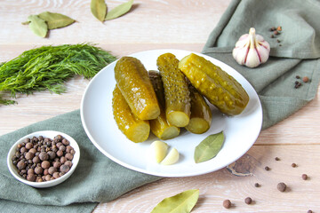 Plate of pickled homemade cucumbers on wooden rustic background. Marinated pickled cucumbers on the white plate with garlic, black pepper and bay leaf. Food concept. Homemade pickled vegetables.