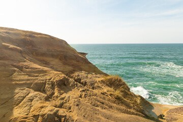 Rocky terrain. Ocean. The water is azure-colored. Light white waves. Clear blue sky. Light clouds. Yellow sand. Beautiful nature. Bright daylight.There is an empty space for your lettering.
