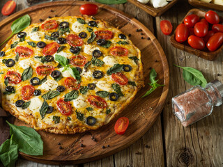 In the photo - pizza on a round wooden dish. Beautiful ruddy crust. Tomatoes, olives, mint leaves. In the background are tomatoes. Rustic background. Dark napkin.