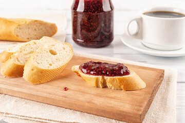 Tasty sandwich with sweet raspberry jam and baguette slices on a wooden cutting board against jam jar and cup of coffe over white wooden table. Breakfast with red berry jelly, marmalade or confiture.