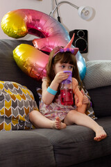 A Cute Baby Girl Sitting On A Sofa With A Colourful Balloon And A Doll
