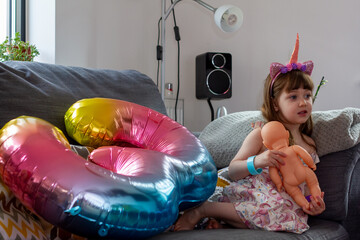 A Cute Baby Girl Sitting On A Sofa With A Colourful Balloon And A Doll