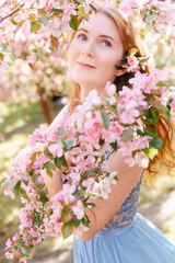 Fototapeta premium Portrait of a young red-haired woman on a background of blooming spring sakura in the park. The concept of fragrant and youthful perfume.