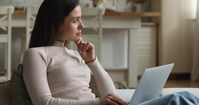 Thoughtful dreamy young woman sitting on sofa thinking working on computer, ponder makes creative task, do telecommuting work on-line using modern device and internet. Creative freelance job concept