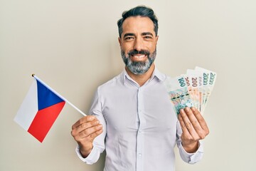 Middle age man with beard and grey hair holding czech republic flag and koruna banknotes smiling with a happy and cool smile on face. showing teeth.