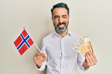 Middle age man with beard and grey hair holding norway flag and krone banknotes winking looking at...