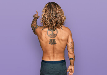 Handsome man with beard and long hair standing shirtless showing tattoos posing backwards pointing...