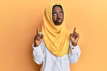 Beautiful african young woman wearing doctor uniform and hijab pointing up looking sad and upset, indicating direction with fingers, unhappy and depressed.