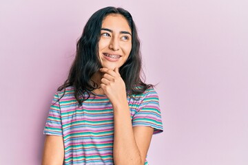 Hispanic teenager girl with dental braces wearing casual clothes with hand on chin thinking about...