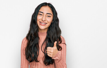 Hispanic teenager girl with dental braces wearing casual clothes doing happy thumbs up gesture with hand. approving expression looking at the camera showing success. - obrazy, fototapety, plakaty