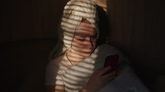 Close up of young woman with towel on her head browsing smartphone in dark room. Portrait of female with mobile phone illuminated by daylight from window through blinds