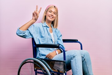 Beautiful blonde woman sitting on wheelchair smiling looking to the camera showing fingers doing...
