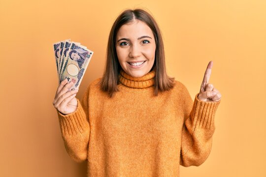 Young woman holding 5000 japanese yen banknotes smiling with an idea or question pointing finger with happy face, number one