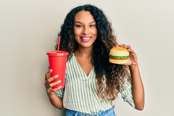 Young latin woman eating a tasty classic burger and soda smiling with a happy and cool smile on face. showing teeth.