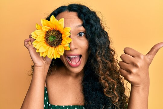 Young latin woman holding sunflower over eye pointing thumb up to the side smiling happy with open mouth