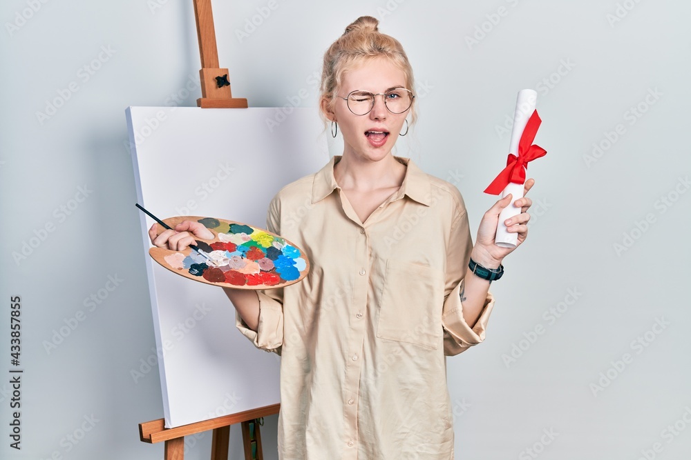 Poster beautiful caucasian woman with blond hair standing by painter easel stand holding diploma winking lo