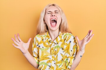 Beautiful caucasian woman with blond hair wearing colorful shirt crazy and mad shouting and yelling...