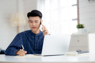 Fototapeta premium Young asian man talking phone and work from home with laptop computer, freelance male writing on notebook and speak on smartphone, stay home, domestic life, business and communication concept.