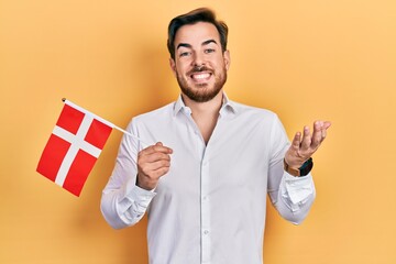 Handsome caucasian man with beard holding denmark flag celebrating achievement with happy smile and winner expression with raised hand