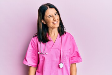 Middle age brunette woman wearing doctor uniform and stethoscope winking looking at the camera with...