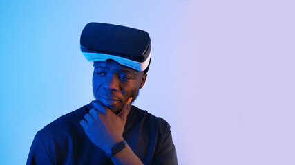 African-American guy with virtual reality glasses on his forehead looking around. Thinking something. Virtual headset glasses. Blue background. Studio footage. . High quality photo