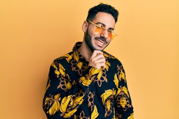 Young hispanic man wearing stylish sunglasses smiling looking confident at the camera with crossed arms and hand on chin. thinking positive.