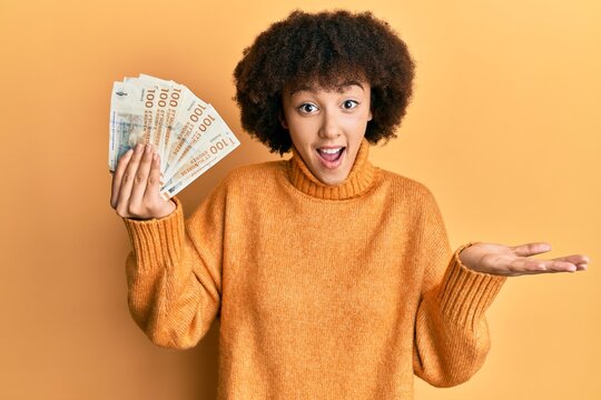 Young hispanic girl holding 100 danish krone banknotes celebrating achievement with happy smile and winner expression with raised hand