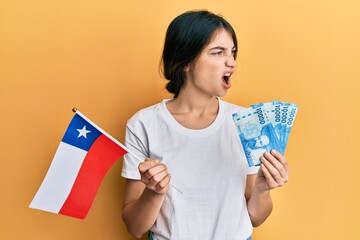 Young caucasian woman holding chile flag and chilean pesos banknotes angry and mad screaming frustrated and furious, shouting with anger. rage and aggressive concept.