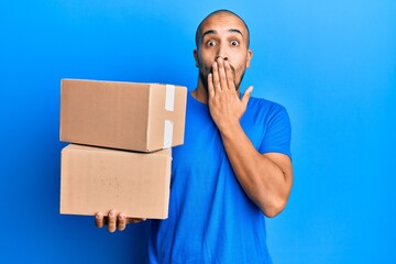 Hispanic adult man holding packages over blue background covering mouth with hand, shocked and...