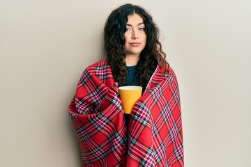 Young brunette woman with curly hair wrapped in a red warm red blanket drinking coffee relaxed with serious expression on face. simple and natural looking at the camera.