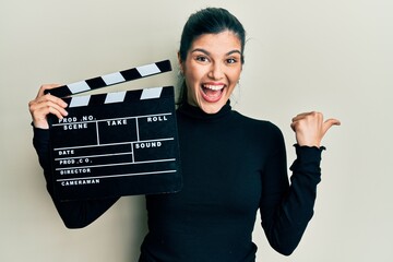 Young hispanic woman holding video film clapboard pointing thumb up to the side smiling happy with...