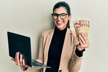 Young hispanic woman wearing business style holding laptop and hungarian forint winking looking at...