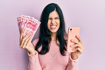 Young hispanic woman using smartphone holding chinese yuan banknotes clueless and confused...